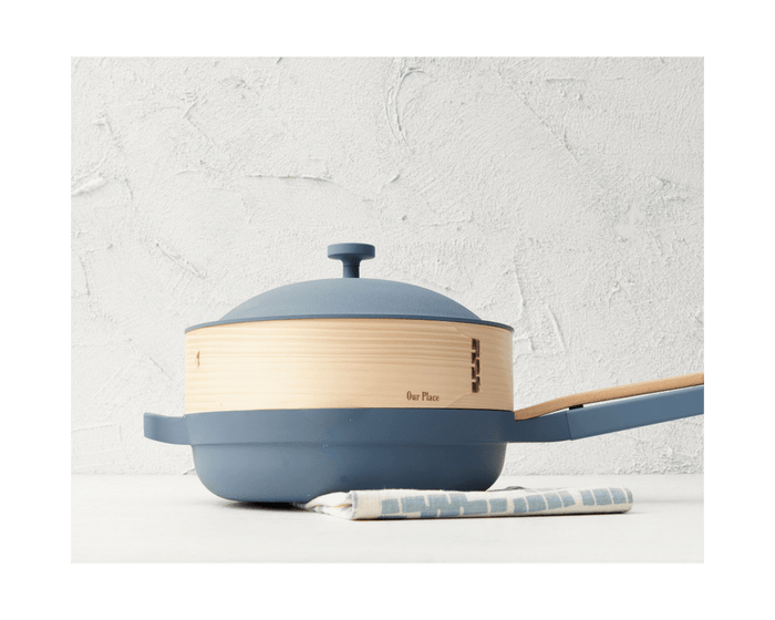 A Spruce Steamer with lid and wooden insert sits on a white surface, bamboo cooking chopsticks resting on a striped cloth beneath the handle.