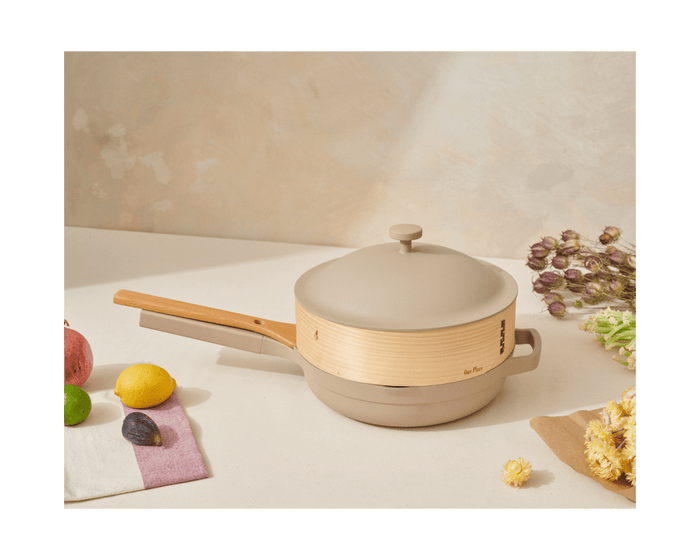 A beige and light wood Spruce Steamer with a lid sits on a countertop next to assorted fruits, a white napkin, and dried flowers.