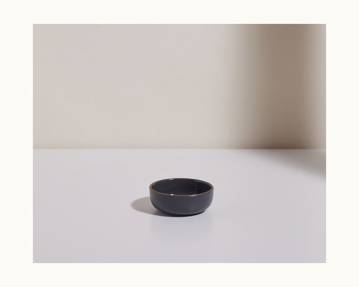 The Tiny Bowls, crafted in dark ceramic, sit on a white surface with a soft right shadow. Their stackable design and scratch-resistant finish add practical style, making them perfect for showcasing elegant tableware against a pale background.