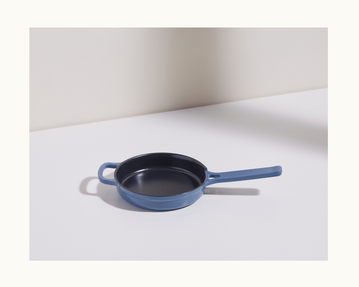 A Tiny Cast Iron Always Pan in blue with two handles is set on a white surface by a light-colored wall.