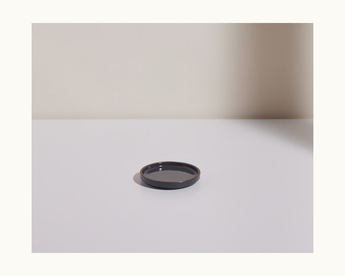 A Tiny Plates small, round, black ceramic plate rests on a white surface in front of a light wall, with a shadow cast on the right.