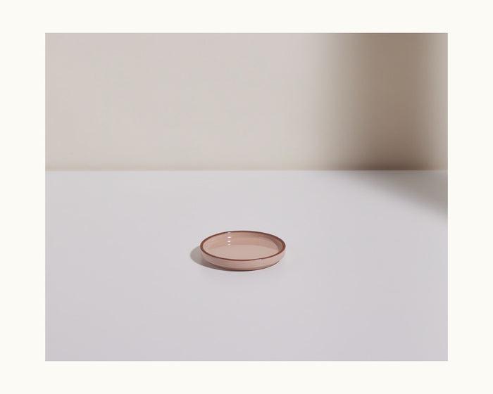 A Tiny Plates snack plate, small and round in pink, is centered on a plain white surface with a soft shadow cast on the wall behind.