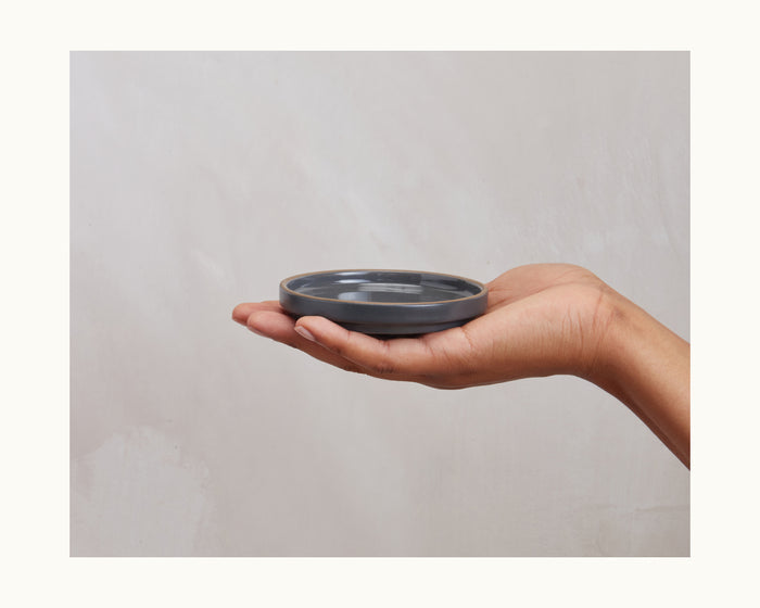 A hand holds a Tiny Plates flat, round, dark gray ceramic plate against a plain light background.