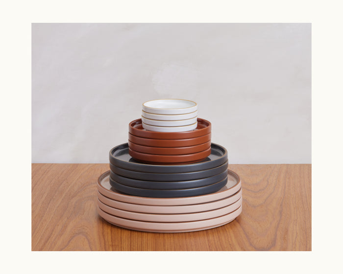 Tiny Plates are stacked in pink, dark grey, and brown, arranged by size on a wooden table to create a stylish tableware set against a light background.