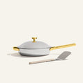 The Titanium always pan pro non toxic frying pan, a white no-coating nonstick frying pan with a gold handle, sits on a light surface with a metal spatula placed in front.