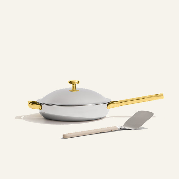 The Titanium always pan pro non toxic frying pan, a white no-coating nonstick frying pan with a gold handle, sits on a light surface with a metal spatula placed in front.